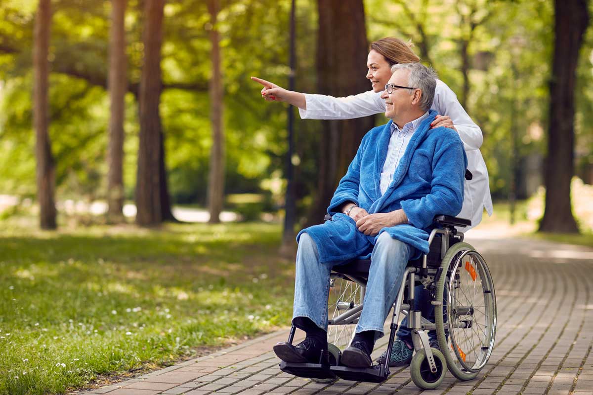 elderly man on hospice smiling and being are being pushed in wheelchair around the park by his caregiver