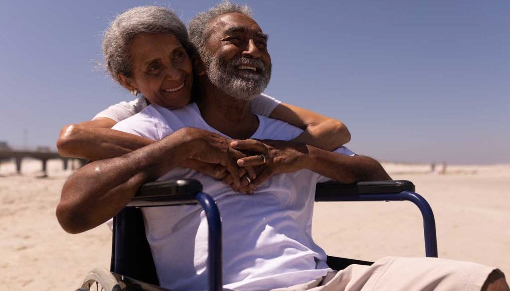 smiling man in hospice care using a wheelchair with his wife's arms wrapped around him enjoying the sun at the beach