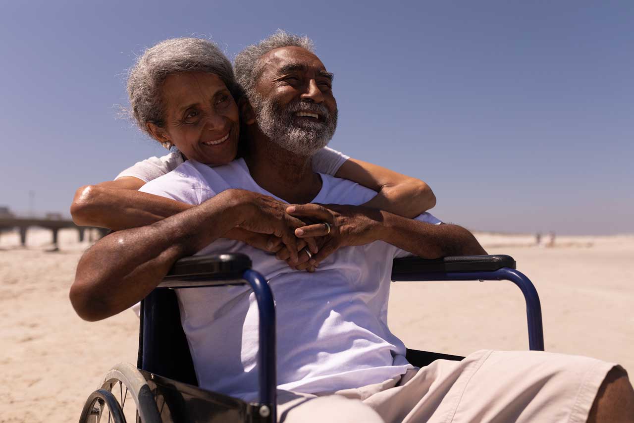 smiling man in hospice care using a wheelchair with his wife's arms wrapped around him enjoying the sun at the beach